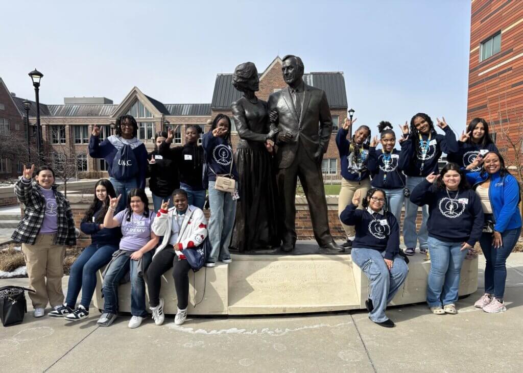 A group of students pose and make peace signs in front of a statue of two people shaking hands on a university campus.