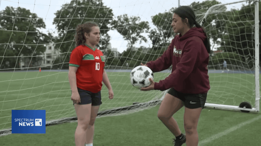 NY1 Scholar Athlete Dahna, in a maroon hoodie, hands a soccer ball to a younger student by the goal post