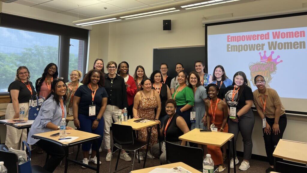 A group of women pose and smile in a classroom at the NYSACAC conference, with a screen behind them displaying Empowered Women Empower Women.