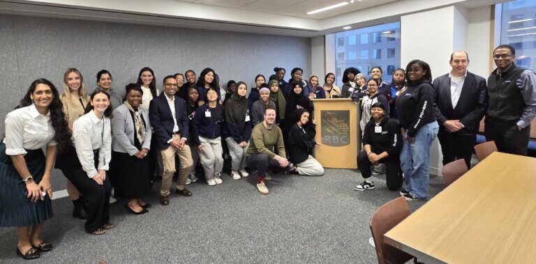 A large group of people pose for a photo in a conference room, some standing and some kneeling, with an RBC podium