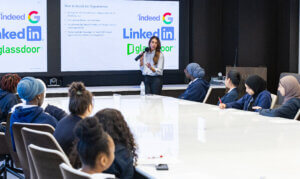 A woman stands and speaks in front of a large screen displaying job search websites to a group of students seated around a conference table.