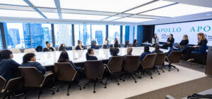 A group of people sits around a large conference table in a modern office, listening to speakers in front of a screen displaying “APOLLO.”.