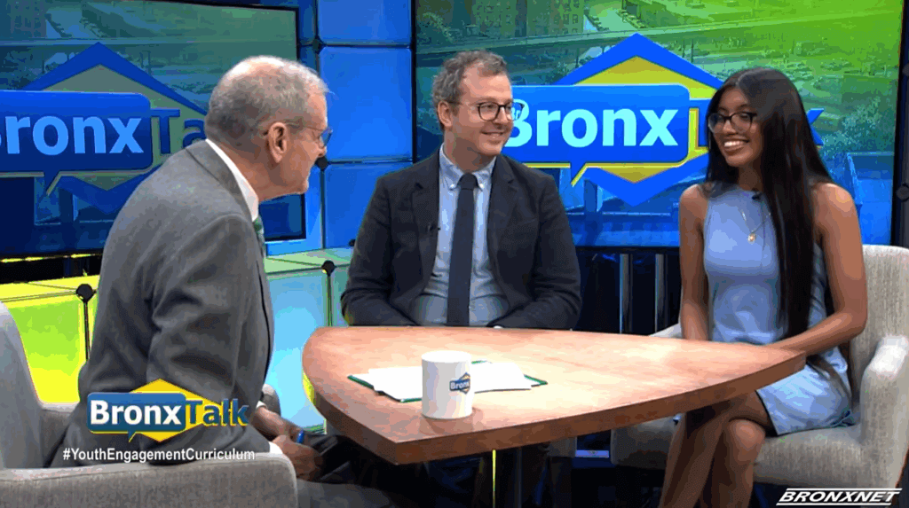 Student and two adults sit at a round table on the set of BronxTalk, discussing student civic engagement