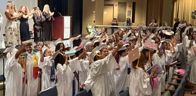 A group of graduating students in white gowns stands and raises decorated caps while people on stage applaud during a graduation ceremony in an auditorium.