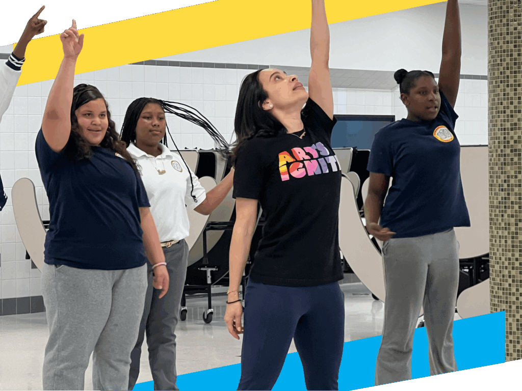Four people stand indoors, three in uniform and one in a black ARTS IGNITE t-shirt, all raising one arm upward in a group activity.