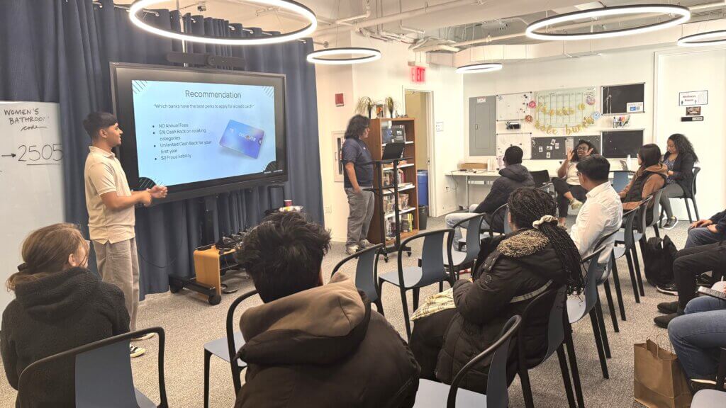 A group of students sits in a classroom, listening to two presenters standing by a screen displaying a slides about personal finance for students