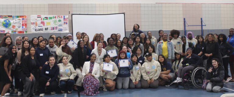 A large group of people posing indoors, some holding certificates, in front of a screen and decorated wall banners.