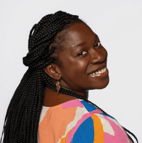 A woman with long braided hair smiles over her shoulder, wearing a colorful patterned top and gold earrings, poses for WILMA Women to Watch Finalists photo shoot