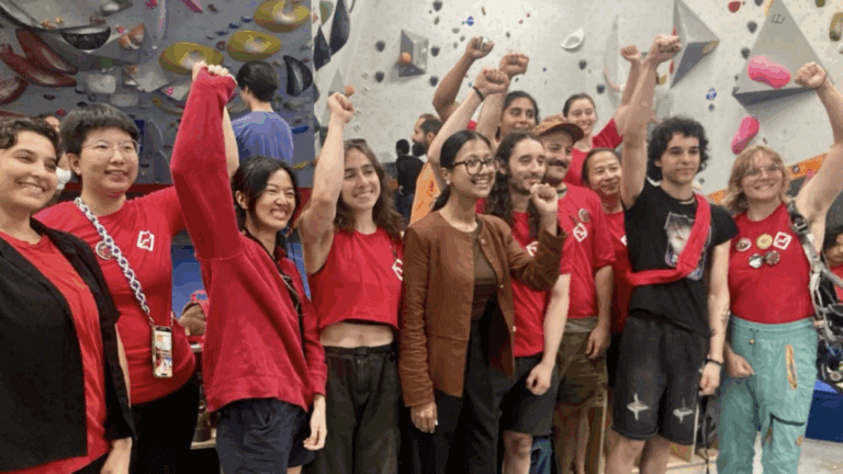 A group of people stand indoors by a rock climbing wall, raising their fists and smiling in front of the climbing wall