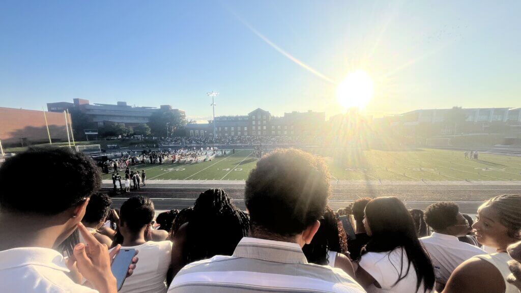 People sit in stadium bleachers on a sunny day, watching a ceremony taking place on a football field at Howard University