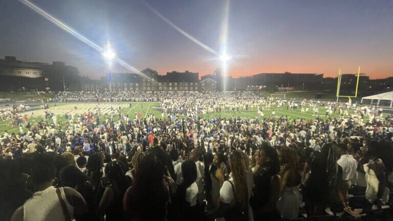 A large crowd gathers on a football field at dusk under bright stadium lights, with spectators filling the stands.