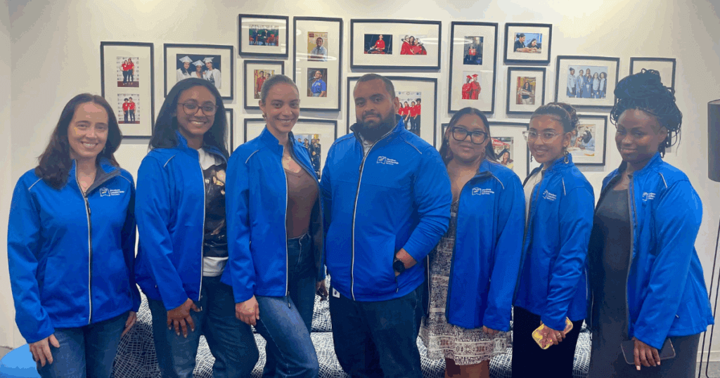 Seven people stand side by side indoors, all wearing matching blue jackets, with framed photos displayed on the wall behind them.