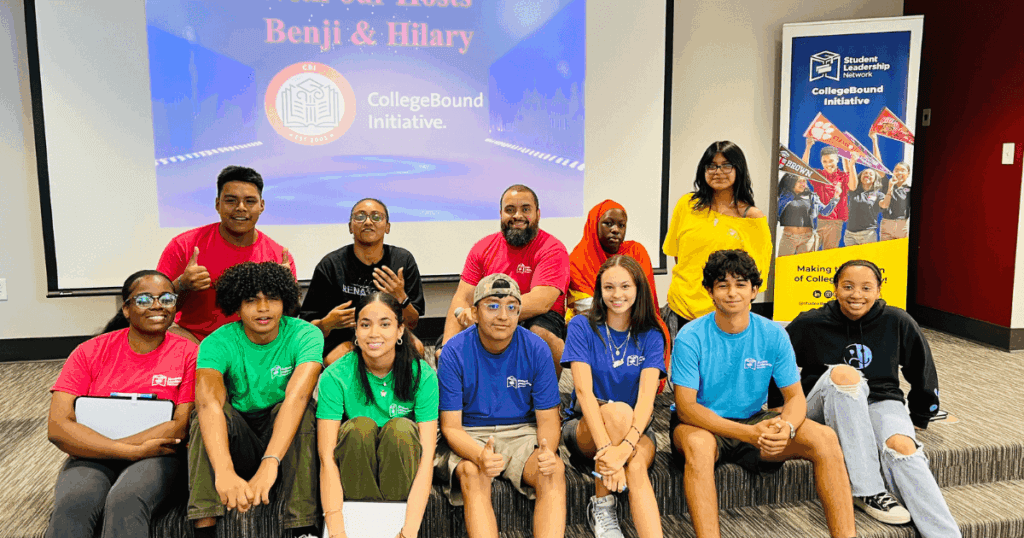 A group of students and adults in colorful shirts sit and stand together in a room with a CollegeBound Initiative presentation on a screen behind them.