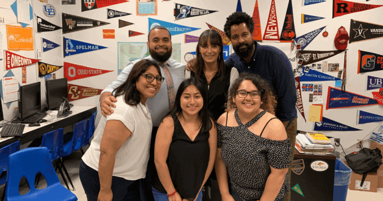 Six adults pose and smile in a classroom decorated with various college pennants on the walls.