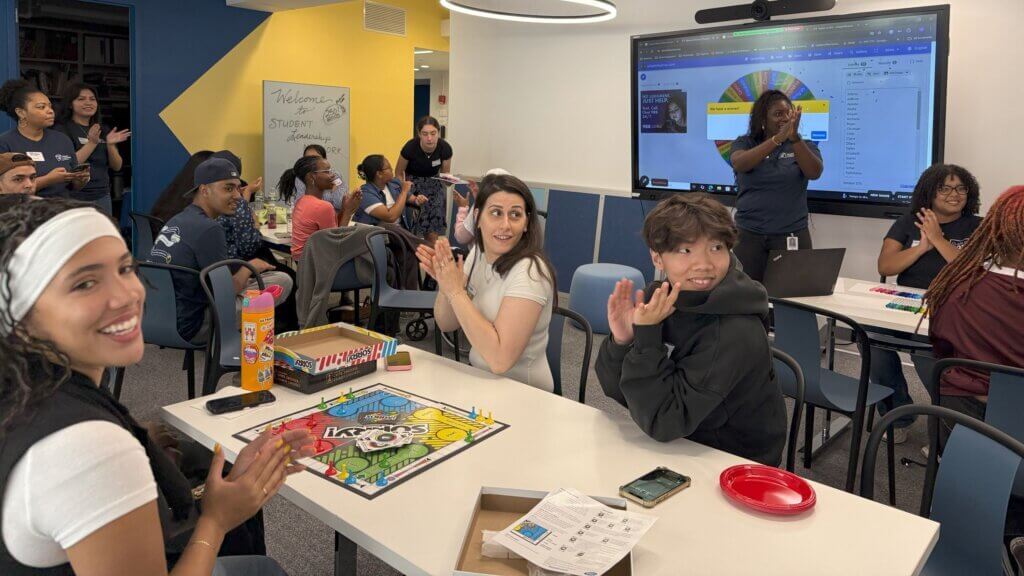 Students sit around tables playing board games and clapping in a classroom, while others watch and a person presents from a screen at the front of the room.