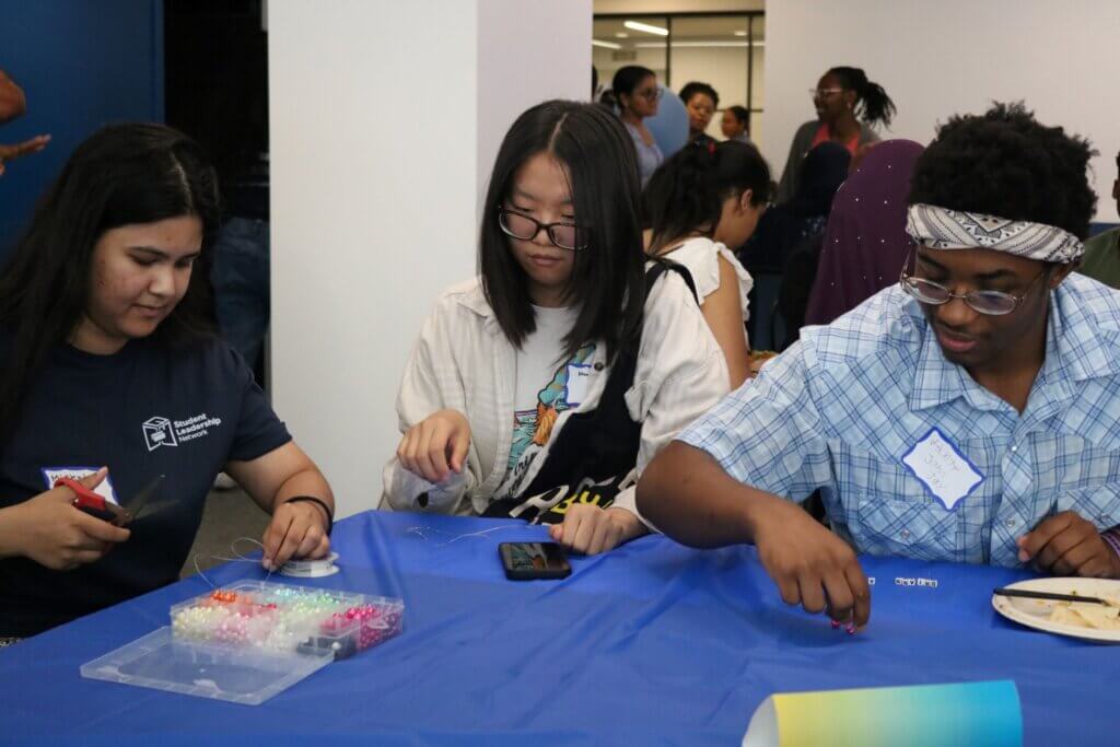 Three young people sit at a table with craft supplies, making bead bracelets
