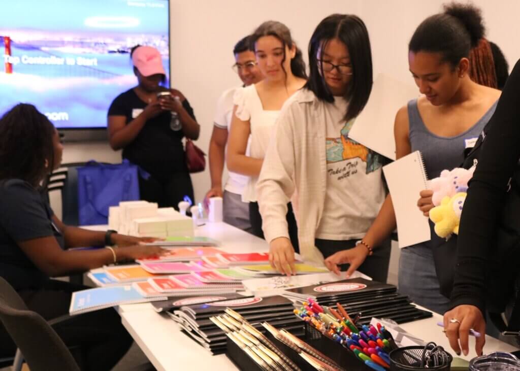 A group of people gather around a table with notebooks, folders, and pens, while a seated person hands out supplies in a brightly lit room.