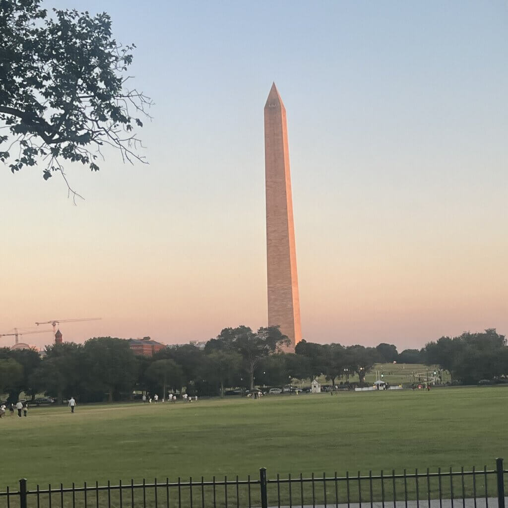 The Washington Monument stands tall at sunset, surrounded by trees and an open grassy area with people walking.