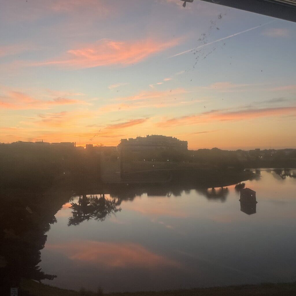 A view outside a dorm window shows a calm sunset over a lake with pink clouds reflecting on the water, buildings in the background.