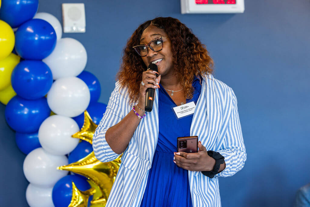 A woman holding a microphone and phone speaks at an event, standing in front of blue, white, and gold balloon decorations.