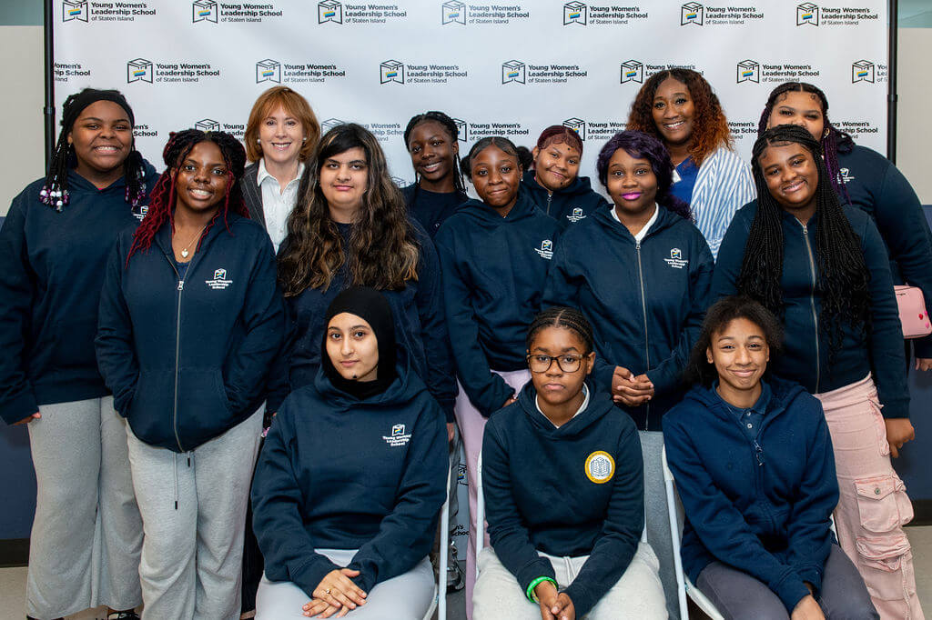 A group of young women and two adults pose together in front of a Young Womens Leadership School step-and-repeat banner at TYWLS Staten Island