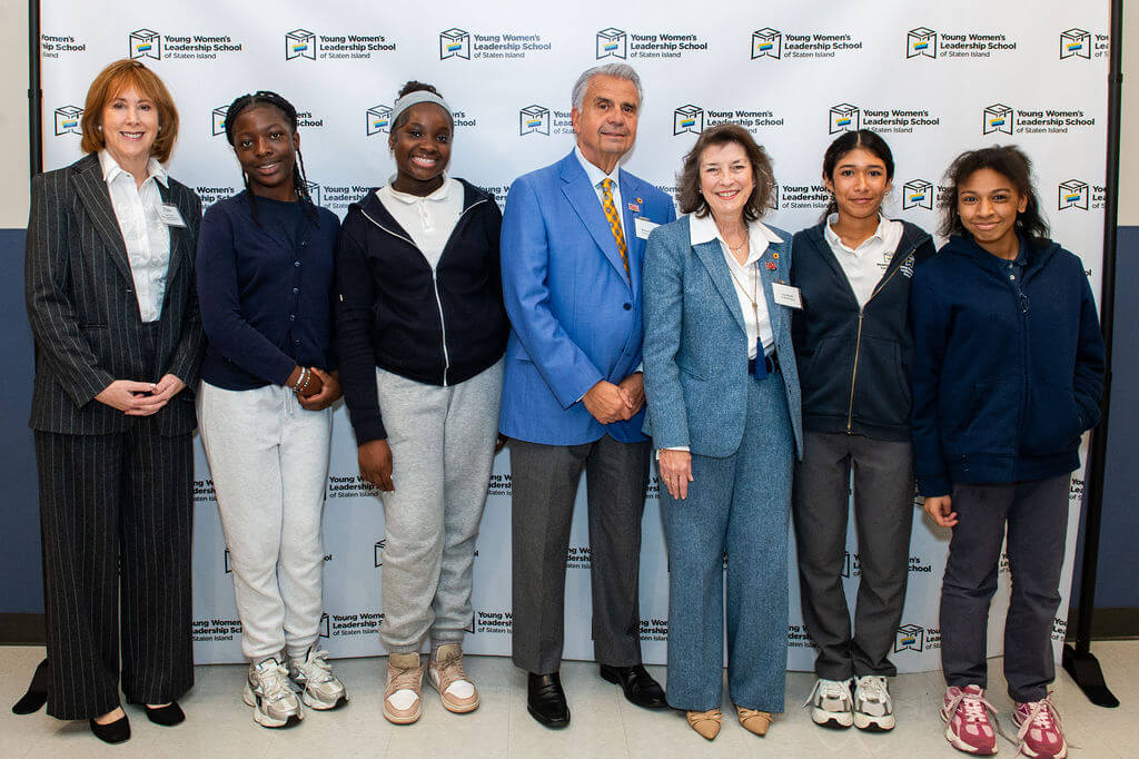 Seven people, including four students in school uniforms and three adults in professional attire, stand in front of a Young Womens Leadership School step-and-repeat banner.