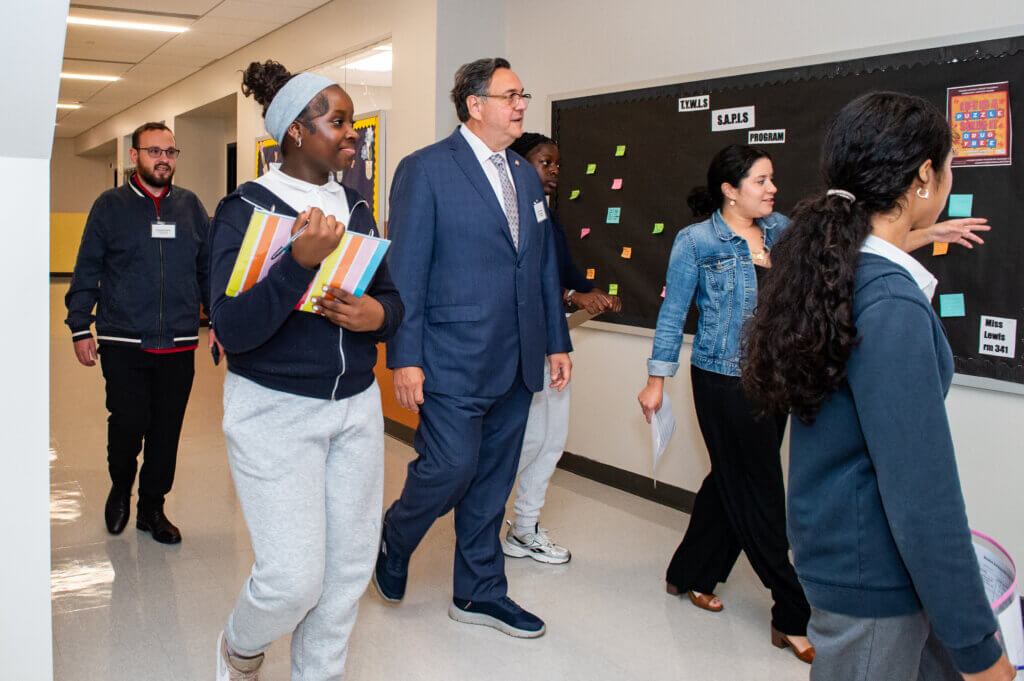 A group of students and adults walk down a school hallway past a bulletin board, some holding notebooks and papers.