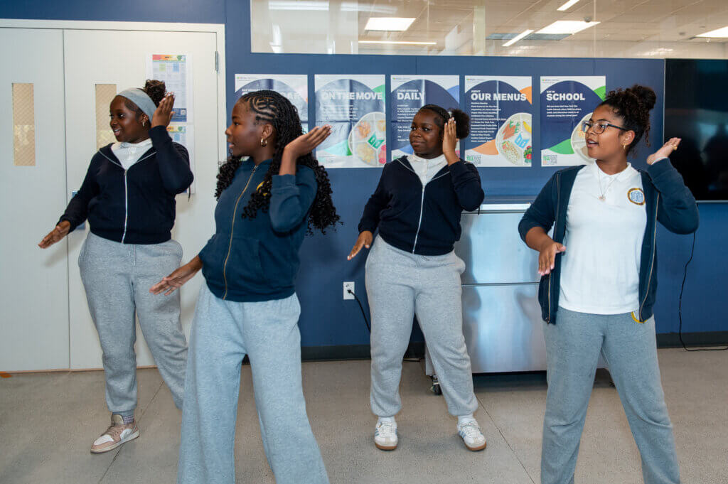 Four students wearing matching school uniforms perform a synchronized dance routine indoors in front of posters on a blue wall.
