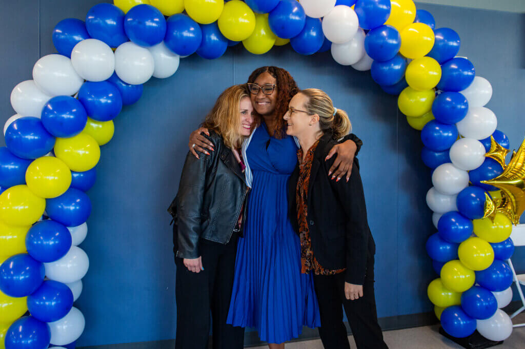 Three women principals hug under a blue, yellow, and white balloon arch at TYWLS Staten Island