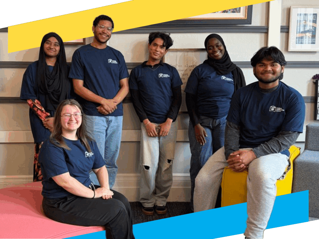 Six college transition coaches wearing matching navy blue shirts pose and smile indoors, sitting and standing on colorful furniture against a decorated wall.