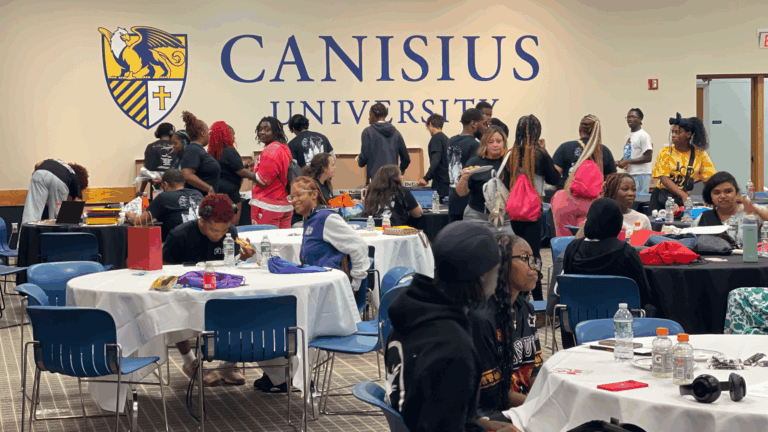 A group of people gather and socialize around round tables in a room with a Canisius University sign on the wall.