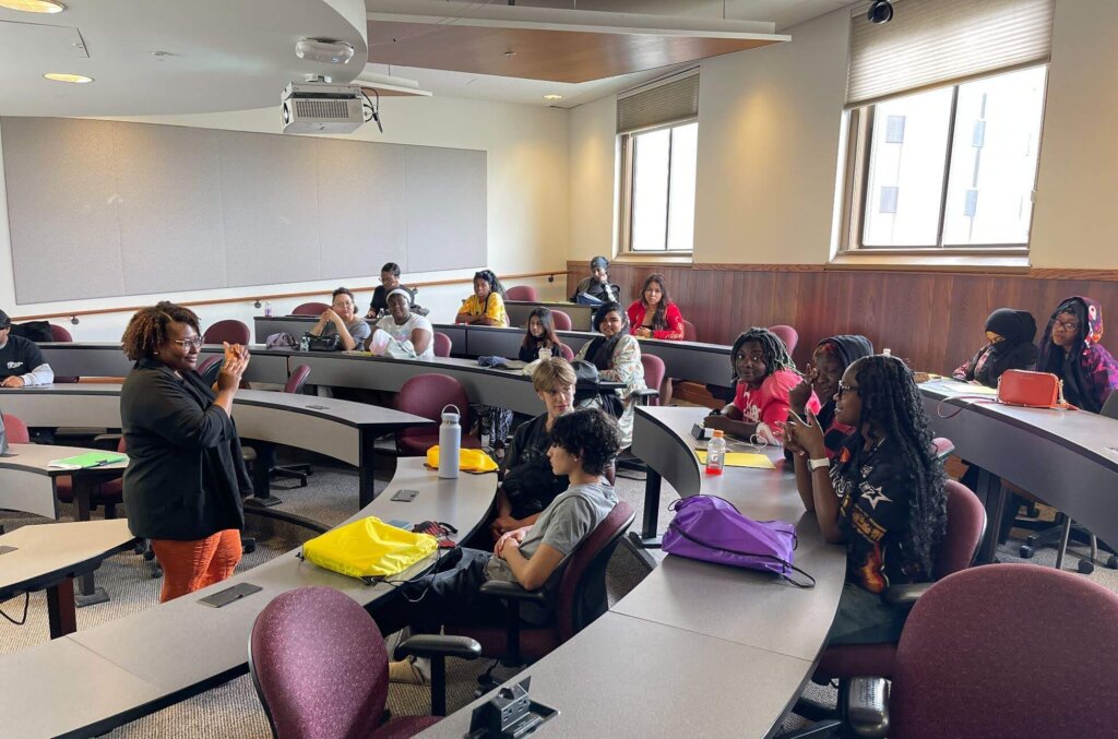 A group of students sit in a curved classroom while a person in front speaks to them; some students take notes and others listen attentively.