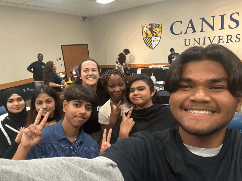 A group of students and an adult smile and pose for a selfie in a classroom with Canisius University on the wall in the background.