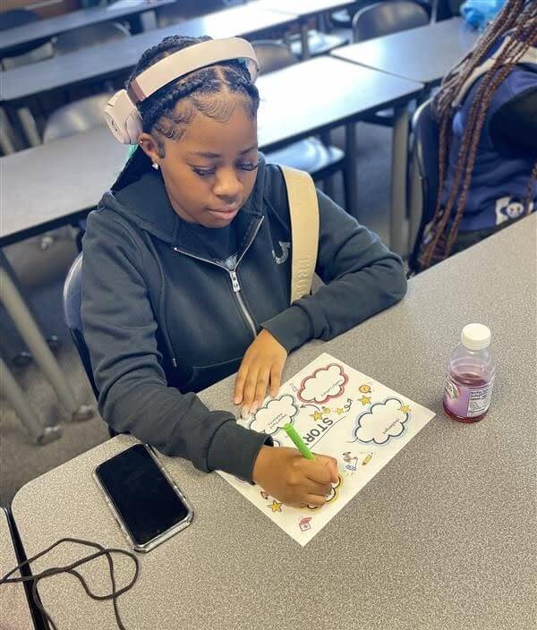 A girl wearing headphones sits at a classroom desk coloring a worksheet with a green marker. A phone and a bottle of juice are on the desk beside her.