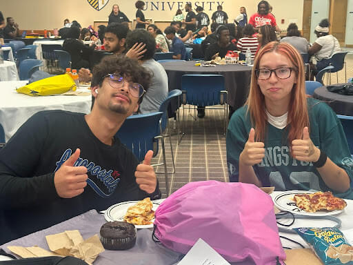 Two students sit at a cafeteria table with food and give thumbs up. Other students are seated at tables in the background.