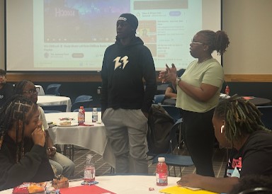 Two people stand and speak to a group seated at round tables in a conference room with a large screen in the background during the Buffalo college access summer program.