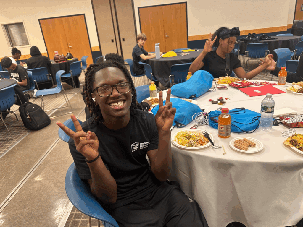 A person in glasses and a black t-shirt sits at a round table with food, smiling and making peace signs during the Buffalo college access summer program. Others nearby enjoy similar meals and drinks, creating a lively, welcoming atmosphere.