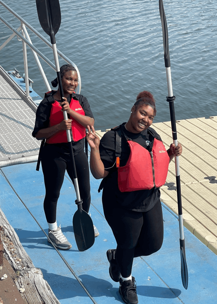 Two young women wearing red life jackets and holding paddles walk up a dock ramp from the water, smiling at the camera.