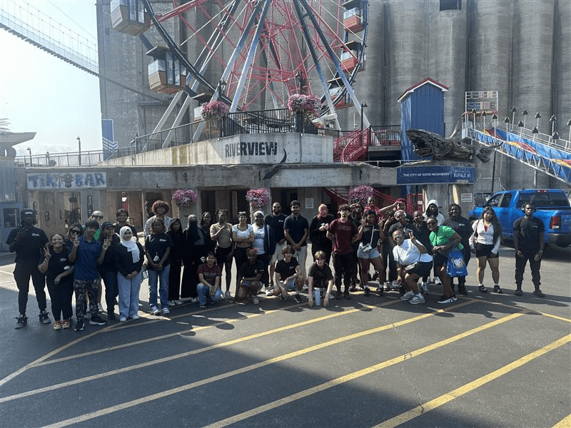 A large group of people poses for a photo in front of a Ferris wheel and a building with Riverview signage on a sunny day.