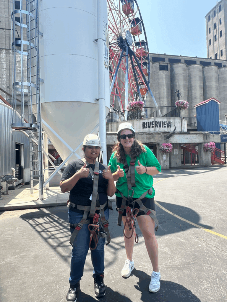 Two people wearing harnesses and helmets stand on pavement, giving thumbs up. A ferris wheel and industrial buildings are visible in the background under bright sunlight.