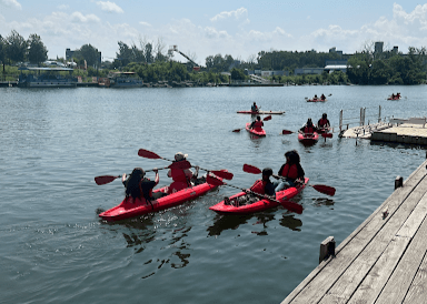 People in red kayaks paddle on a calm river near a wooden dock, with trees and buildings visible in the background under a partly cloudy sky.
