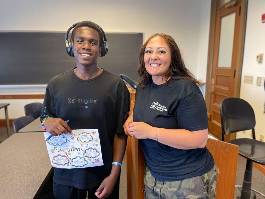 A student wearing headphones holds a colorful story map next to a college counselor at a summer Buffalo college access program.