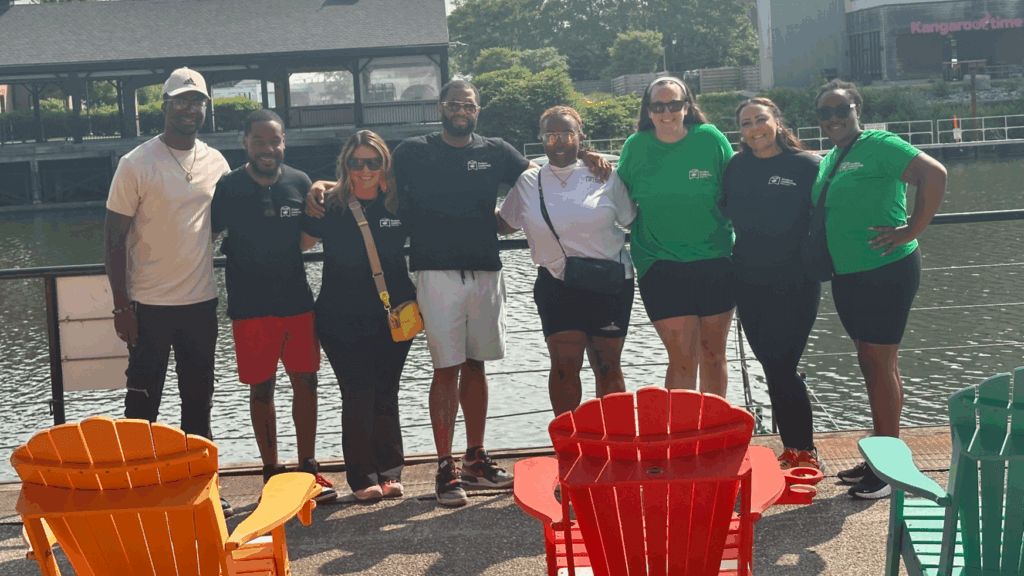 Seven people stand together by a riverside railing, posing for a group photo. Colorful Adirondack chairs face the water in the foreground. Trees and buildings are visible in the background.