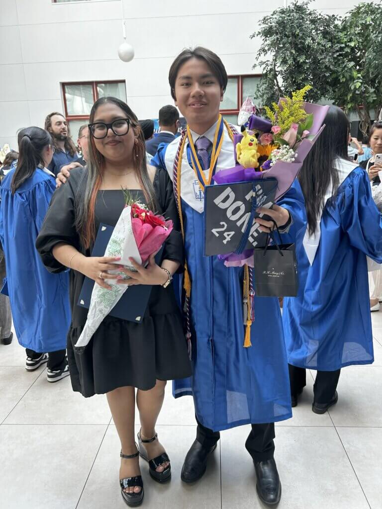 A young man in a graduation cap and gown smiles with his director of college counseling, both are holding flower bouquets
