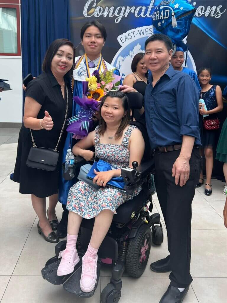 A graduate in a cap and gown stands with three family members, one of whom is in a wheelchair, at a graduation event. Balloons and a congratulatory banner are visible in the background.