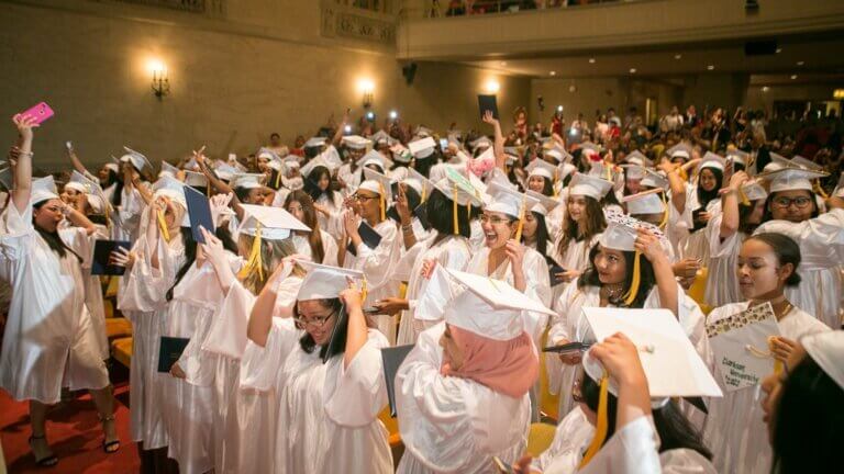 A large group of students in white graduation gowns and caps smile and chat inside an auditorium