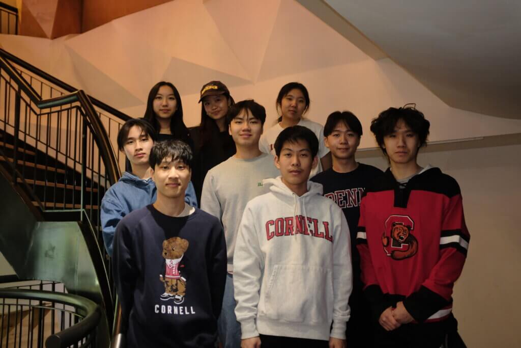 Nine young adults stand on a staircase indoors, some wearing Cornell and Penn sweatshirts, who are part of a college mentoring program