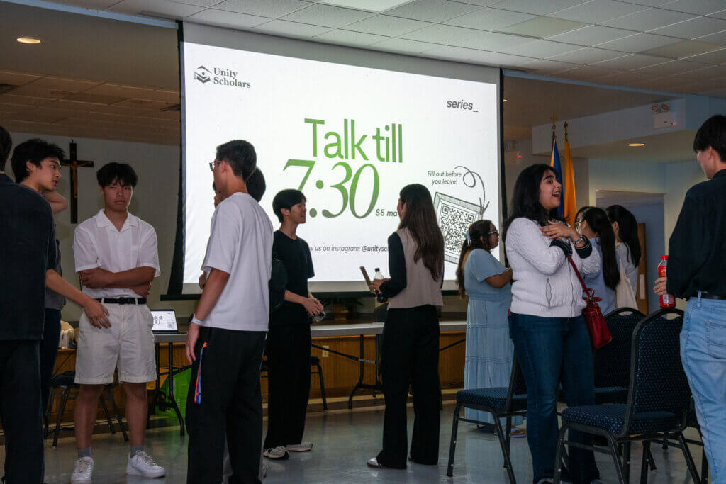 A group of people stand and converse in a room with a large screen displaying Talk till 7:30 and Unity Scholars in the background.