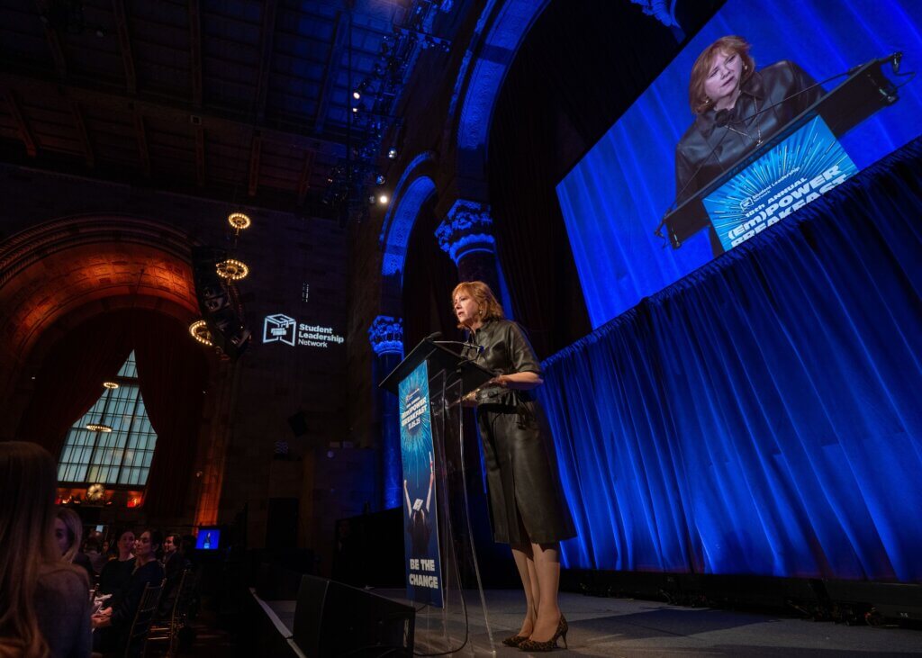 Ann Tisch speaks at a podium on stage, with her image projected on a screen behind her and an audience seated in front.