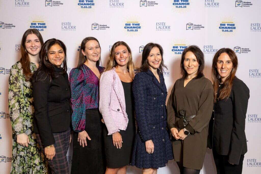 Seven women stand side by side, smiling at the camera, in front of a step-and-repeat backdrop